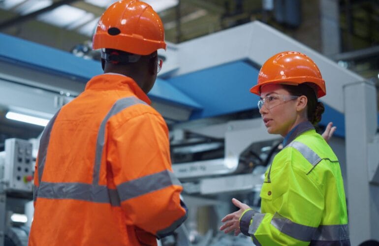 a man and a woman wearing hard harts and high vis, stood in front of automation equipment having a conversation